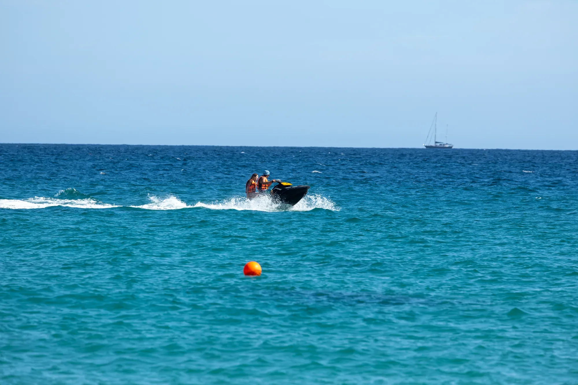 Dos personas en jet ski rumbo a mar abierto con velero al fondo